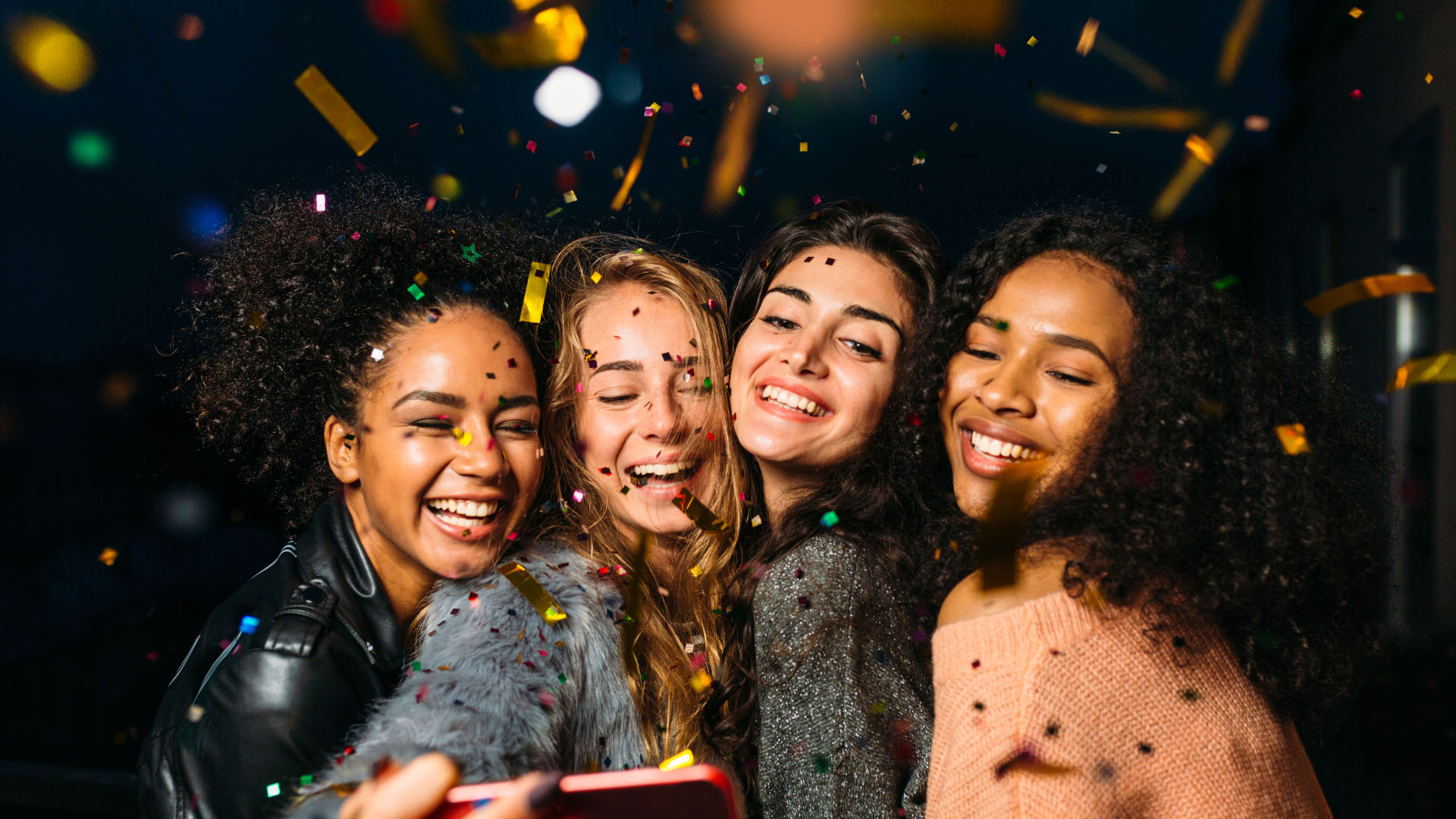 Four laughing women taking a selfie amid falling colourful confetti, displaying bright white smiles at a celebration